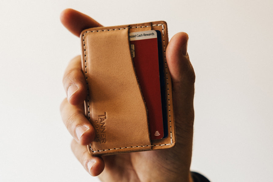 Hand holding tan leather Tanner Goods wallet with red card partially visible, white background, soft lighting.