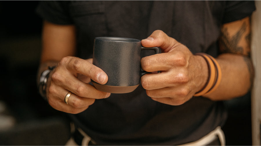 Hands holding a matte black ceramic mug with brown base, wearing rings and orange wristbands, tattooed arm visible.
