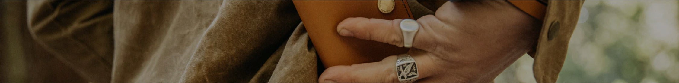 Close-up of hands holding a brown leather bag, wearing silver rings, against a tan coat, soft natural lighting.