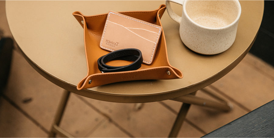 Tan leather tray on round table holds cardholder and black band, beside speckled mug; warm, minimalist aesthetic.