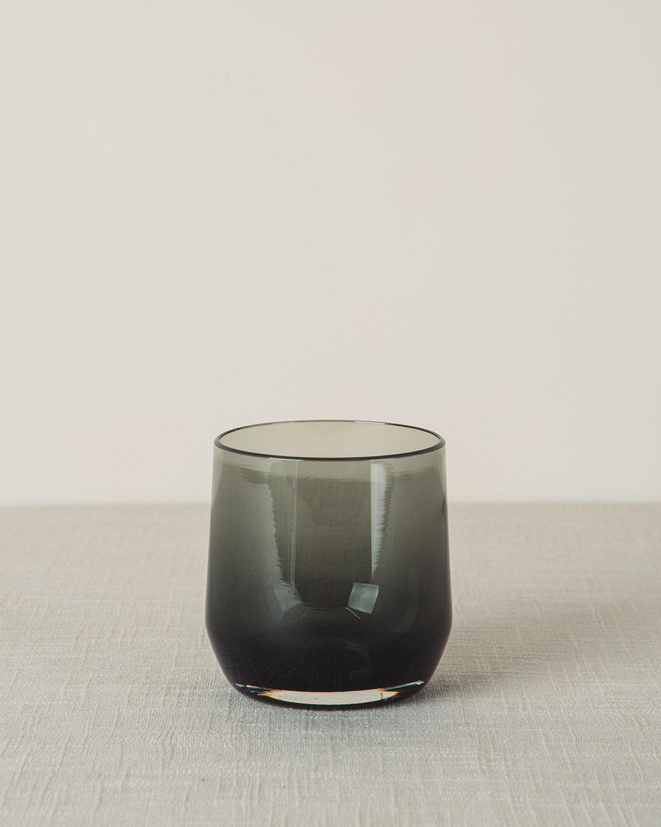 Smoky grey cocktail glass with a modern tumbler shape, resting on a light grey linen table.