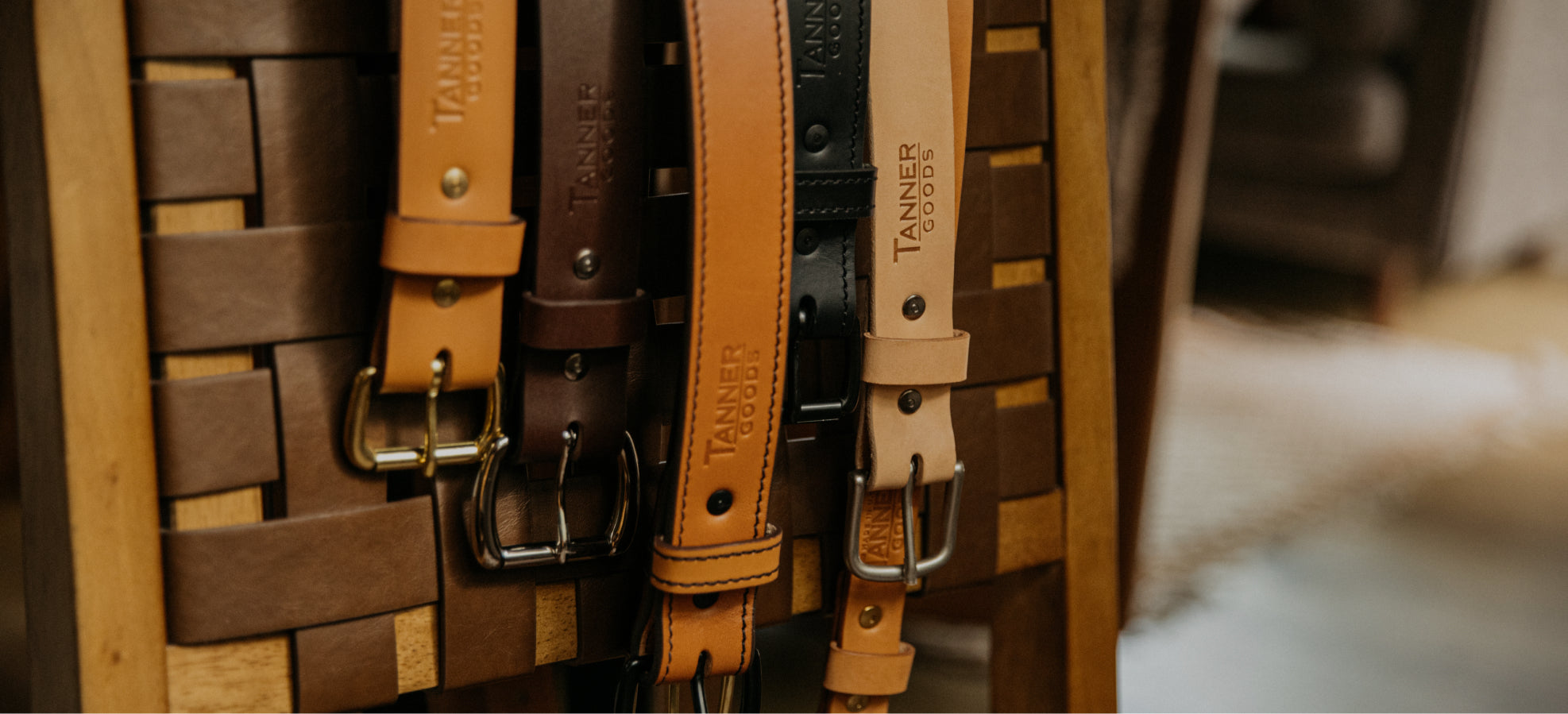 Tanner Goods leather belts in tan, brown, and black with metal buckles, displayed vertically on a woven wooden rack.