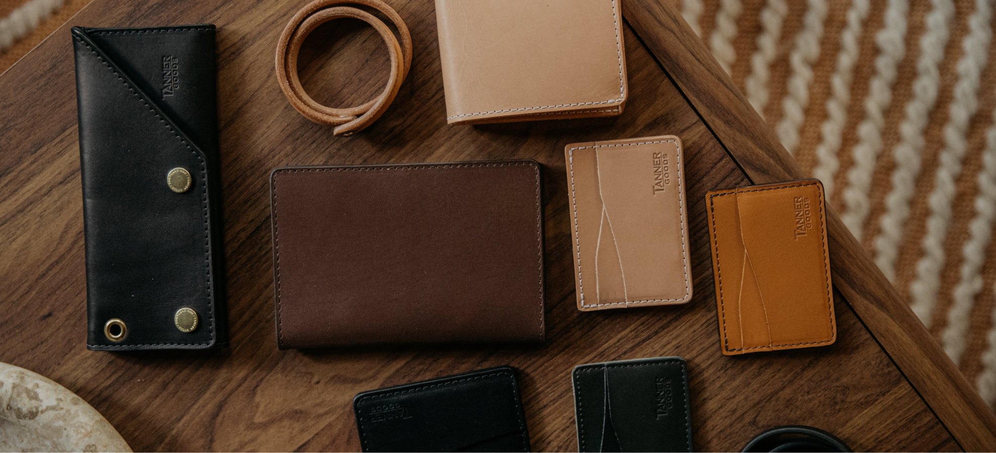 Tanner Goods leather wallets and card holders in black, brown, and tan on a wooden table with a striped rug background.