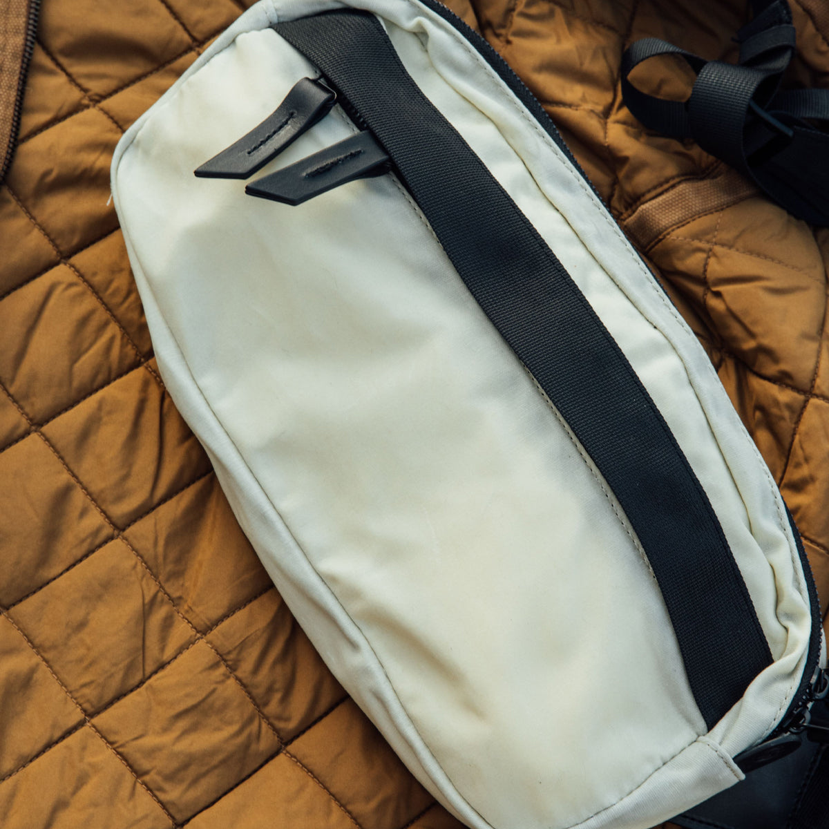 Frost waxed canvas Canyon Crossbody Pack with black webbing and leather zipper pulls, resting on a brown quilted jacket.