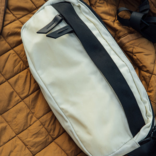 Frost waxed canvas Canyon Crossbody Pack with black webbing and leather zipper pulls, resting on a brown quilted jacket.