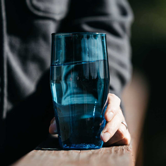 Blue Standard Pint Glass filled with water, held by a hand resting on a wooden railing.