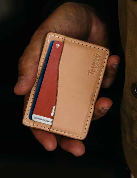 Tan leather cardholder with white stitching, holding red and blue cards, embossed "TANNER GOODS" logo, held in hand against dark background.