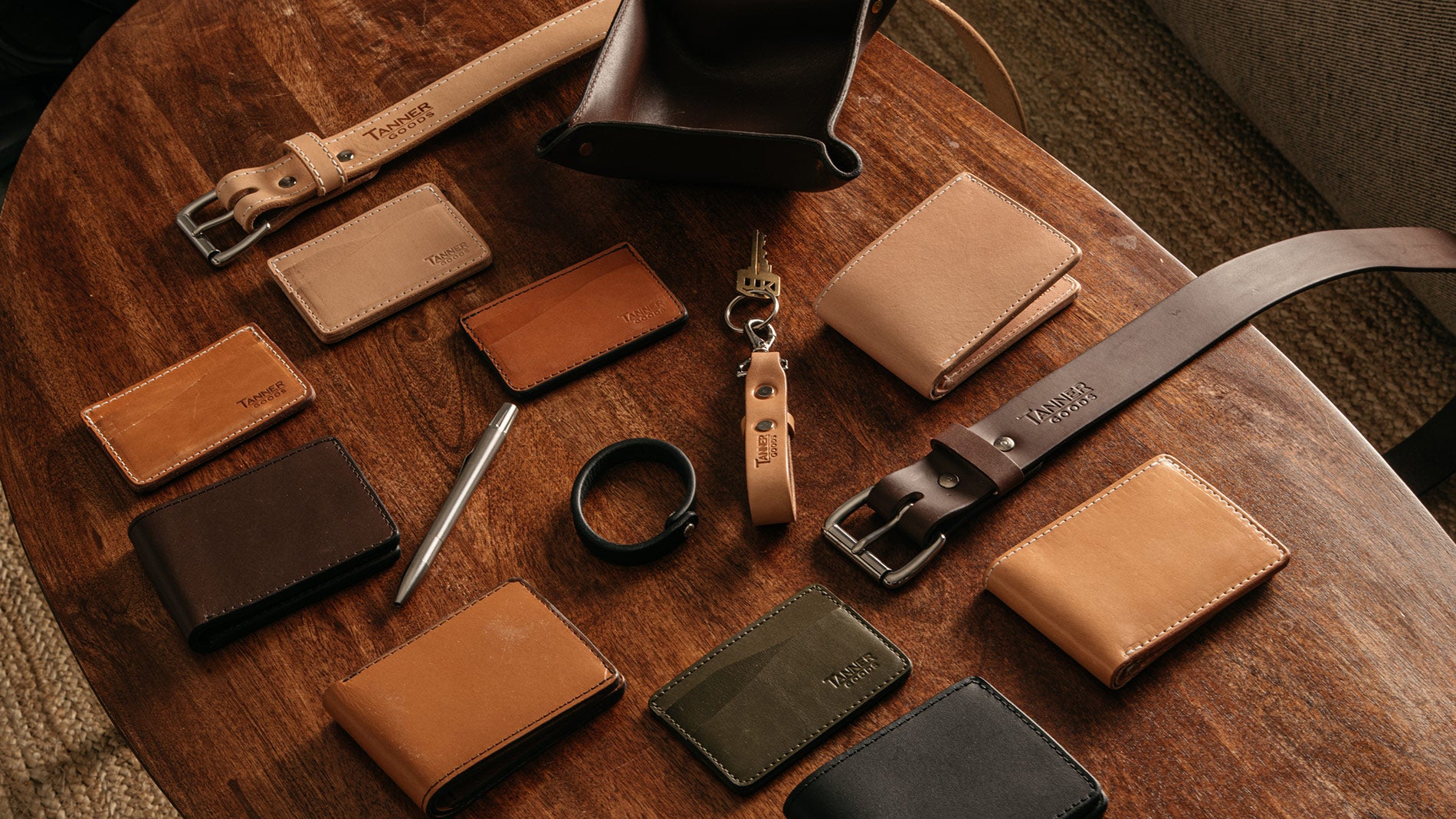 Brown leather wallets, belts, and accessories arranged on a wooden table, branded "Tanner Goods" in warm, natural lighting.