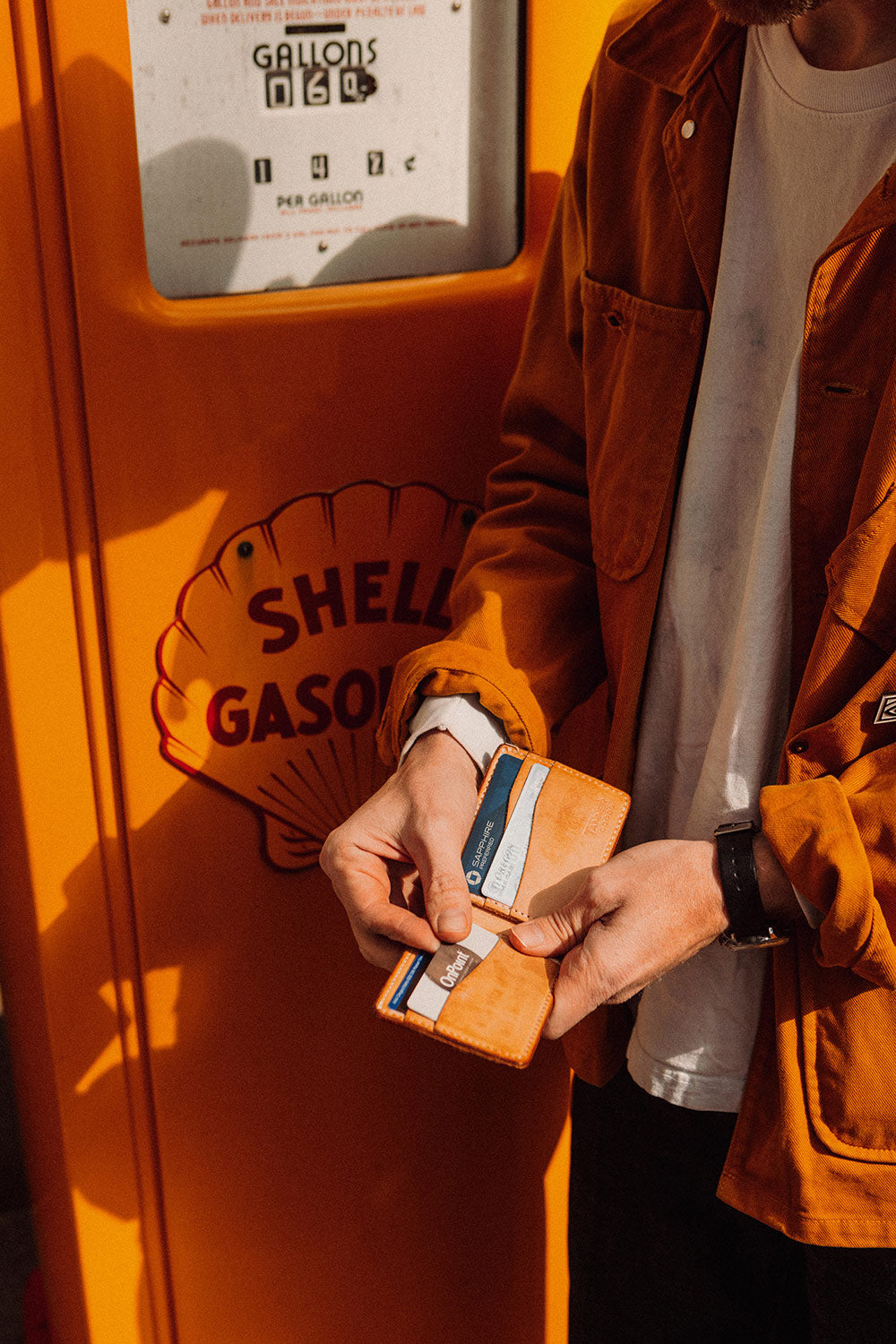 Golden leather bifold wallet held open by hands, showing cards, against an orange Shell gas pump.