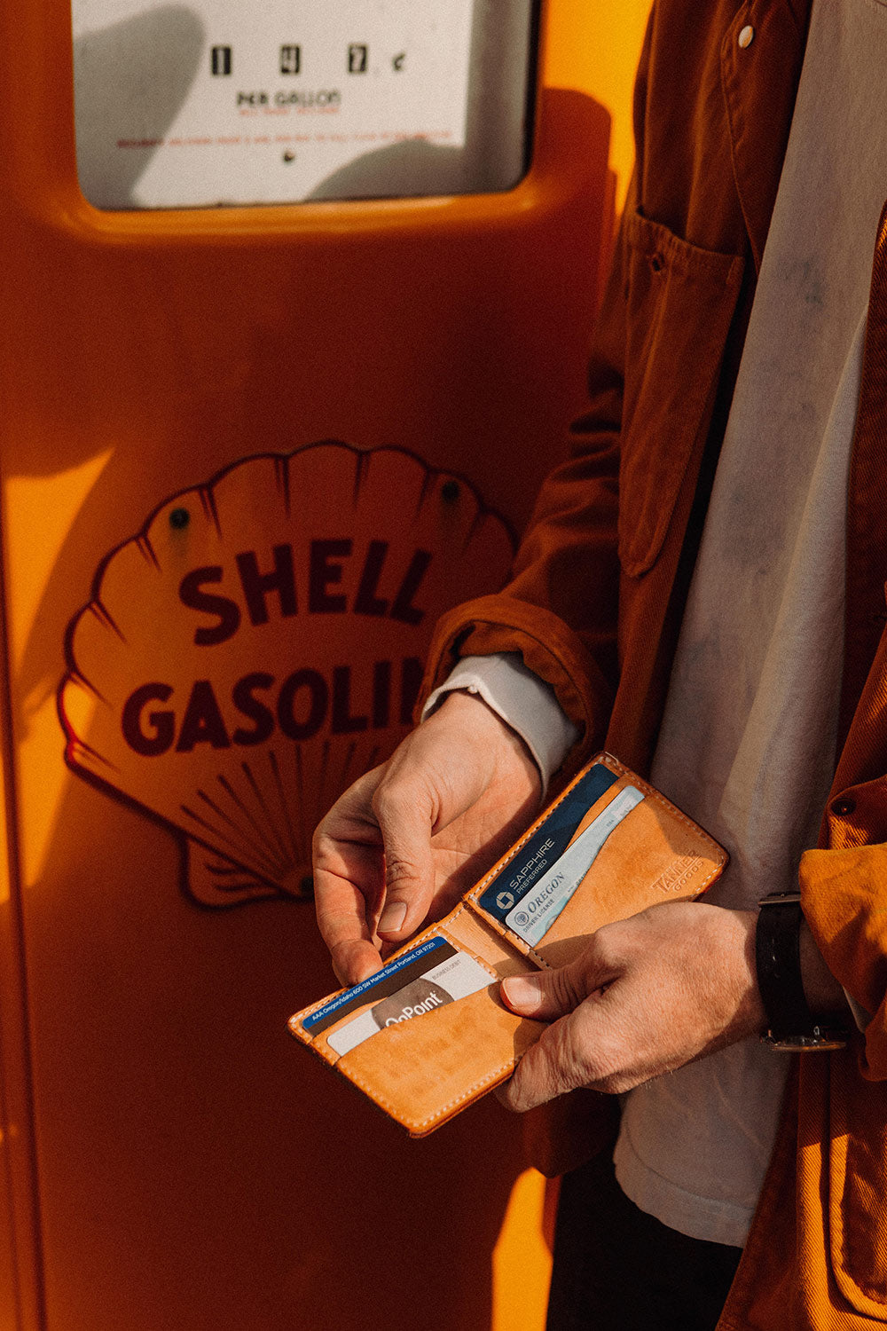 Golden leather bifold wallet held open showing cards, against an orange Shell gas pump.