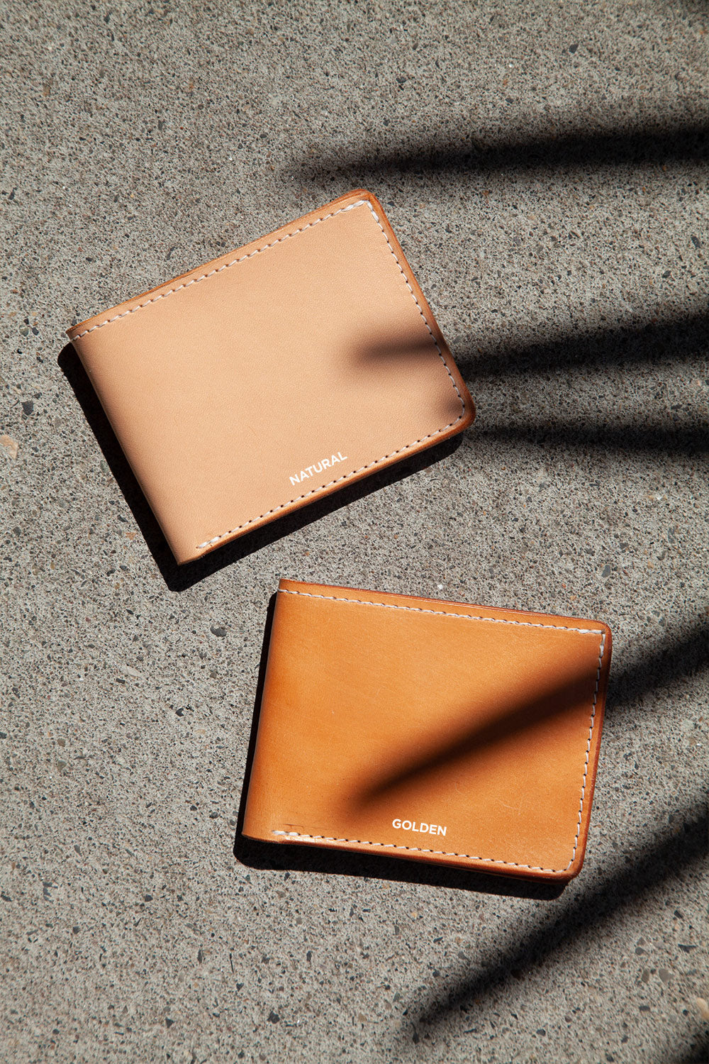 Golden leather bifold wallet with white stitching, beside a natural tan wallet on a concrete surface with shadows.