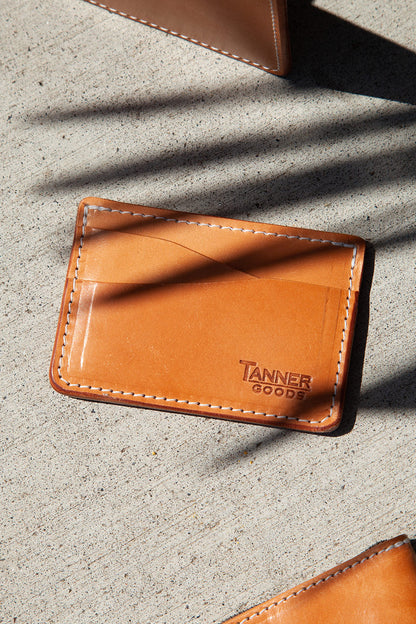 Golden leather wallet by TANNER GOODS with white stitching and wavy card slots, on a concrete surface.