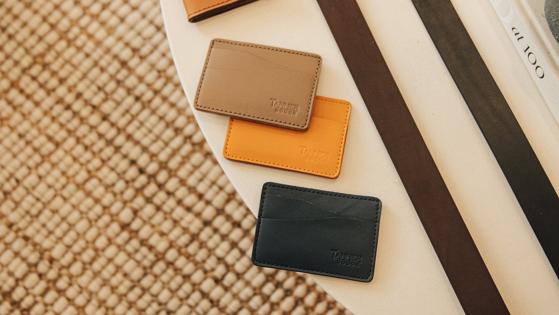 Three Tanner Goods leather card holders in tan, orange, and black arranged on a white surface with woven texture background.