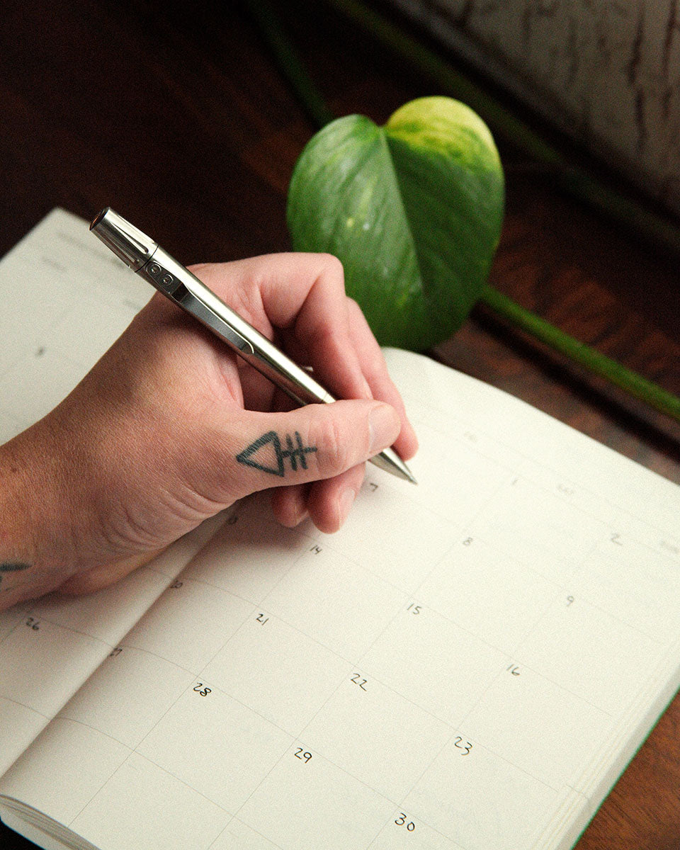 Hand with tattoo writing in calendar notebook, silver pen, green plant nearby, warm wooden surface.