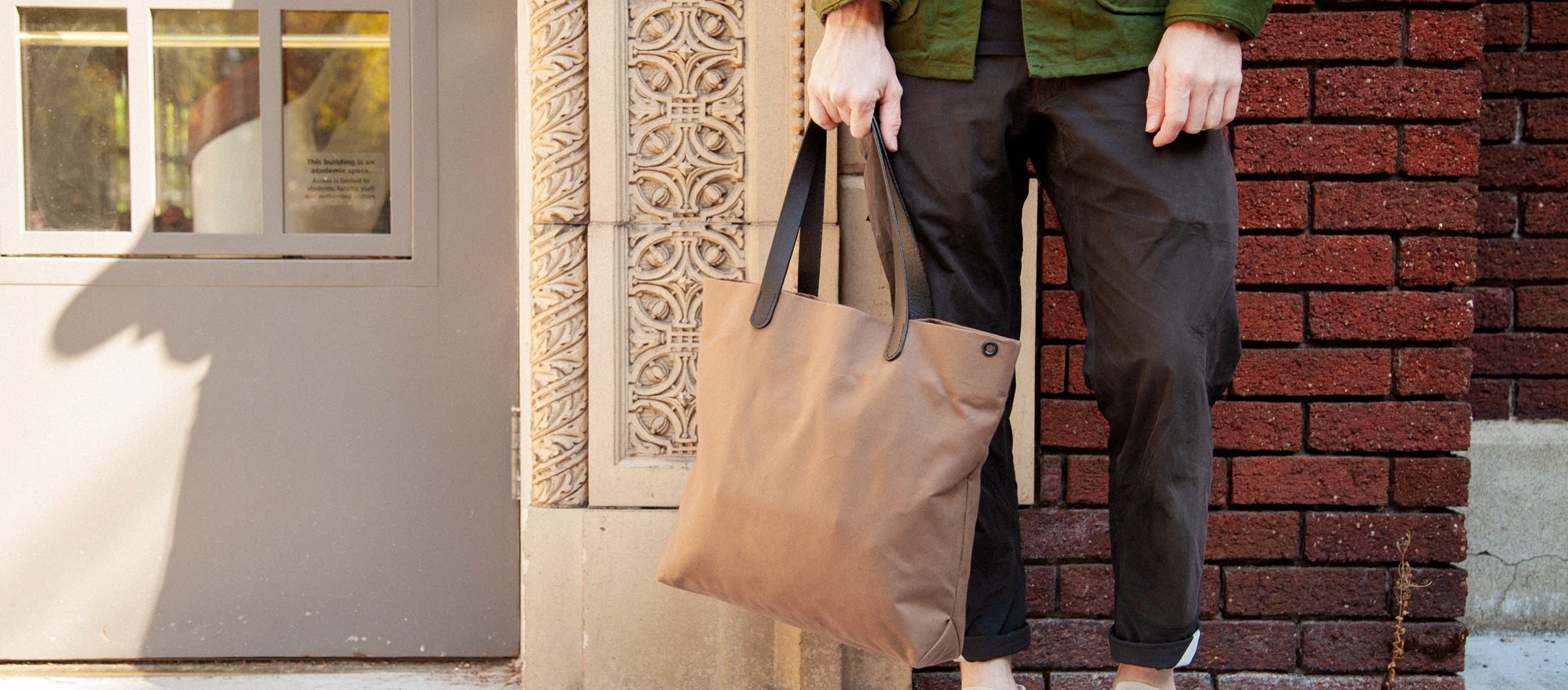 Person in green jacket and black pants holds tan tote bag with black straps, standing beside red brick wall and gray door with ornate stone trim.