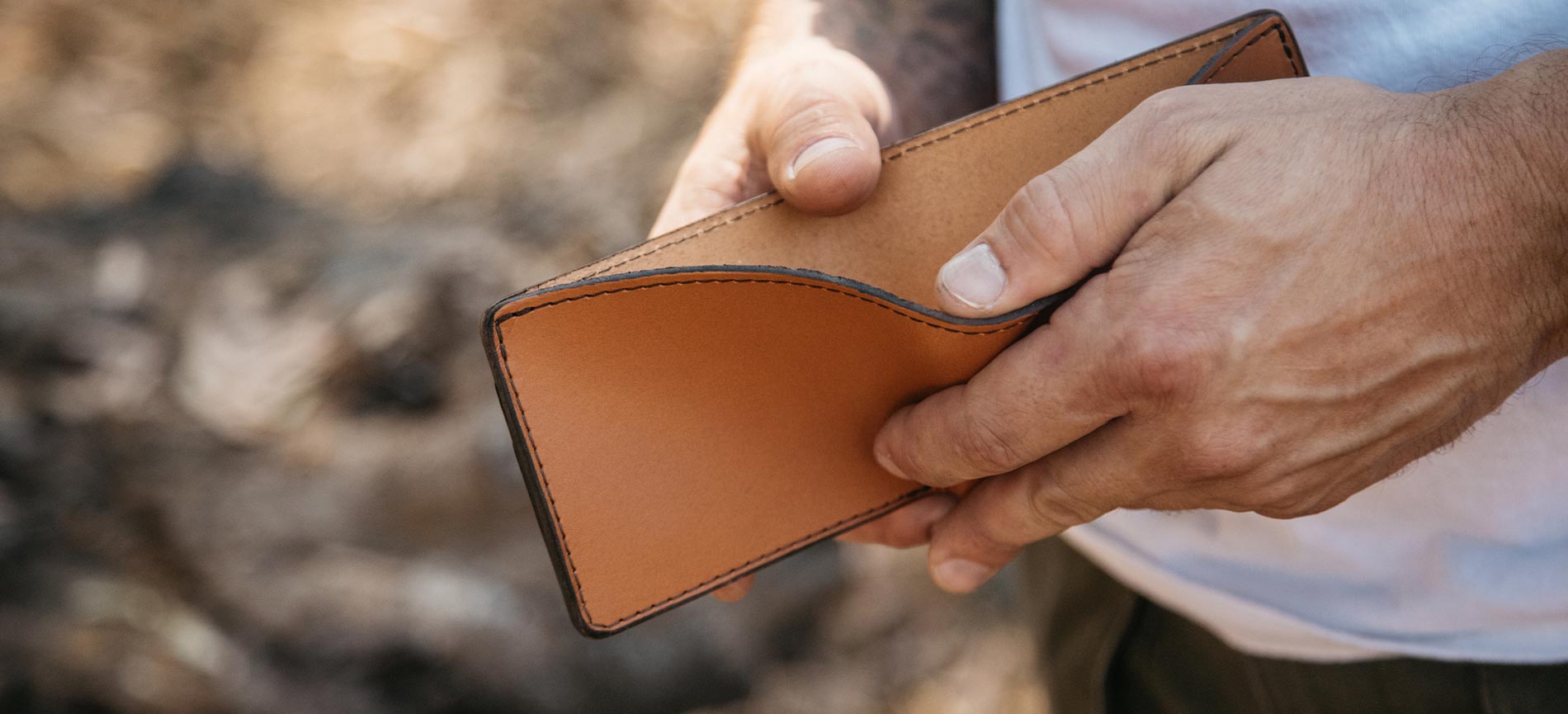 Hands holding a tan leather wallet with black stitching, outdoors, natural lighting.