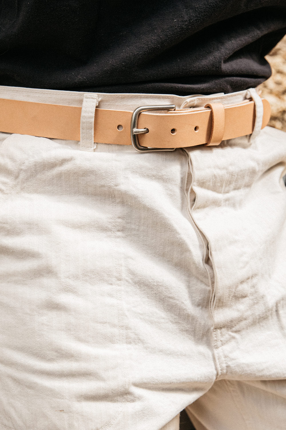 Natural tan leather belt with a silver-toned buckle, worn with light beige pants and a black top.