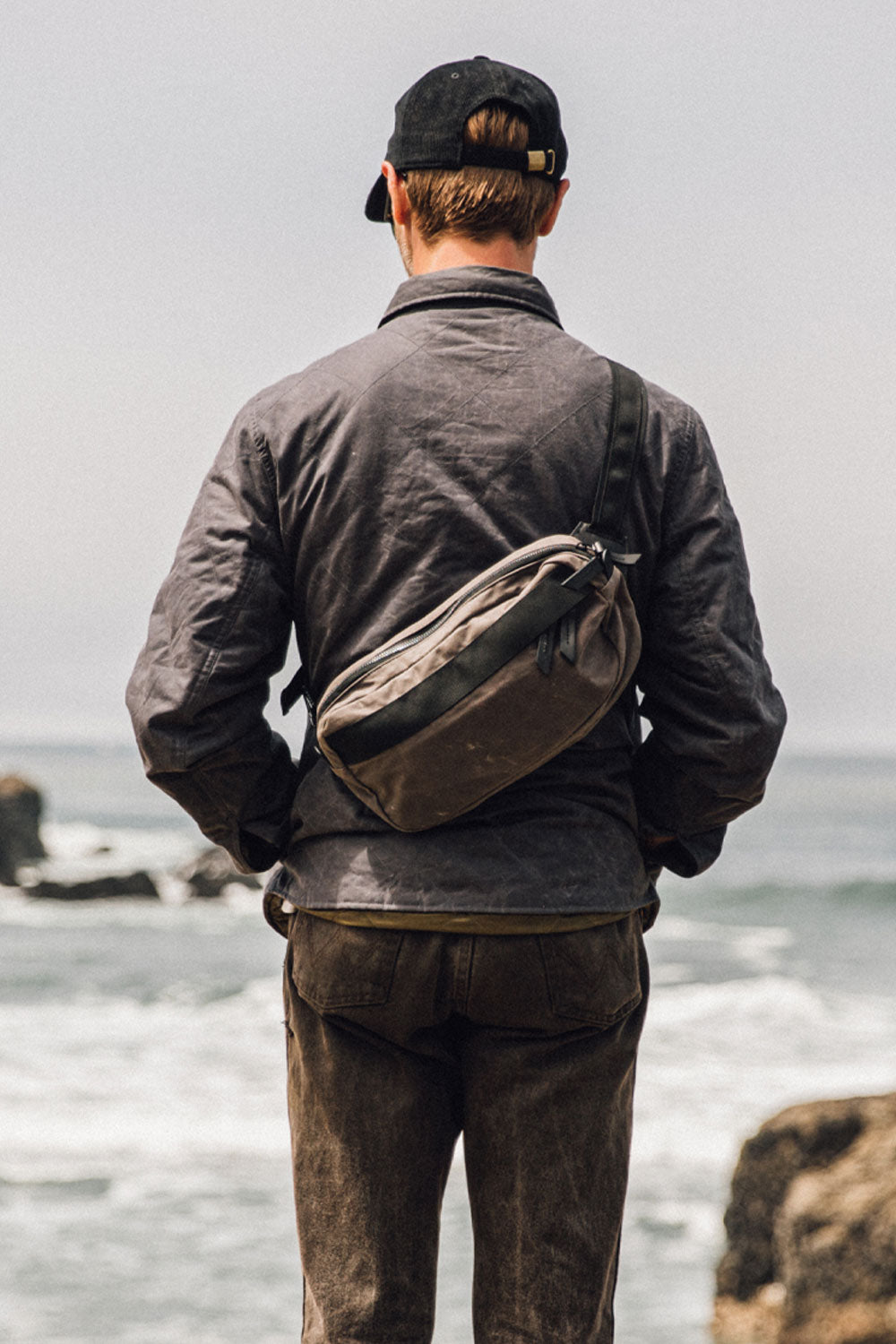 Slate grey waxed canvas crossbody pack worn across the back of a person in a dark jacket by the ocean.
