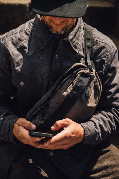 Slate Grey waxed canvas Canyon Crossbody Pack with black zippers and strap, worn across a man's chest as he holds a phone.