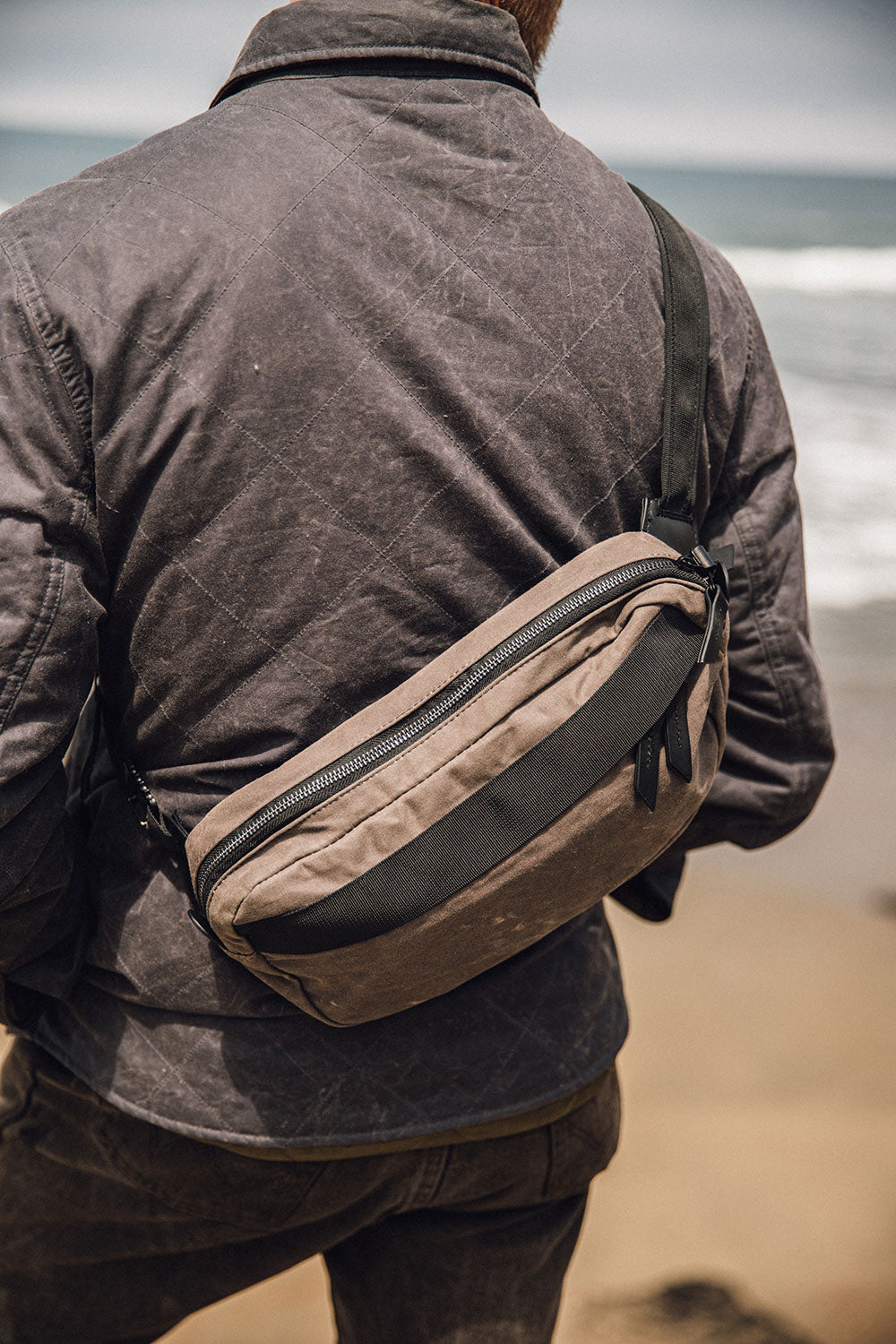 Slate Grey waxed canvas Canyon Crossbody Pack worn across a person's back, wearing a dark quilted jacket by the sea.