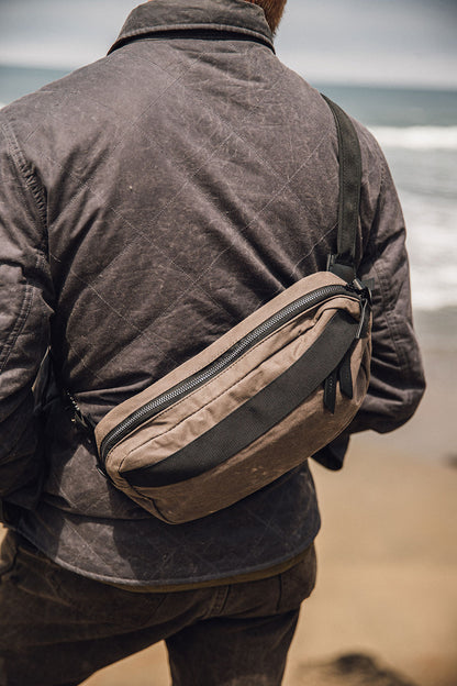 Slate Grey waxed canvas Canyon Crossbody Pack worn across a person's back, wearing a dark quilted jacket by the sea.