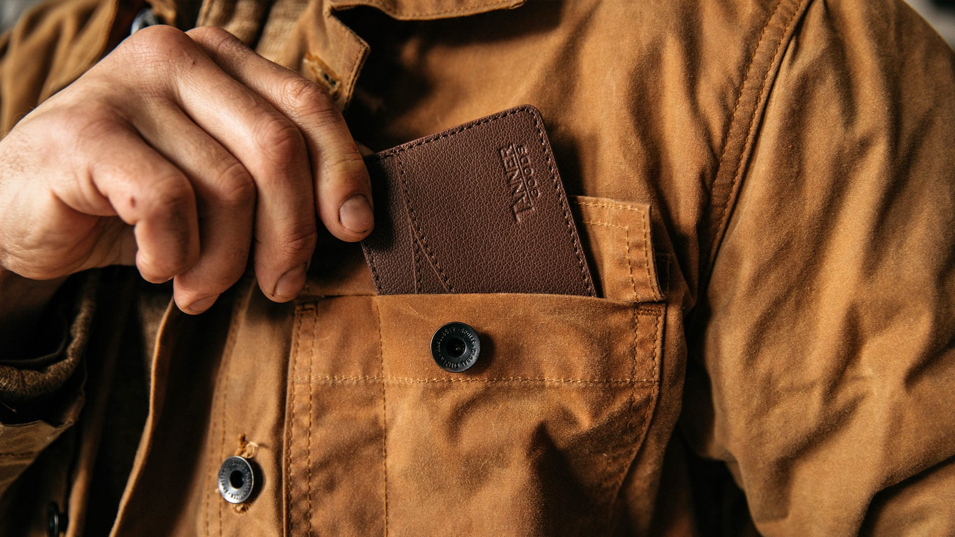 Hand placing brown leather wallet into chest pocket of tan work jacket, embossed "WANDER" logo visible.