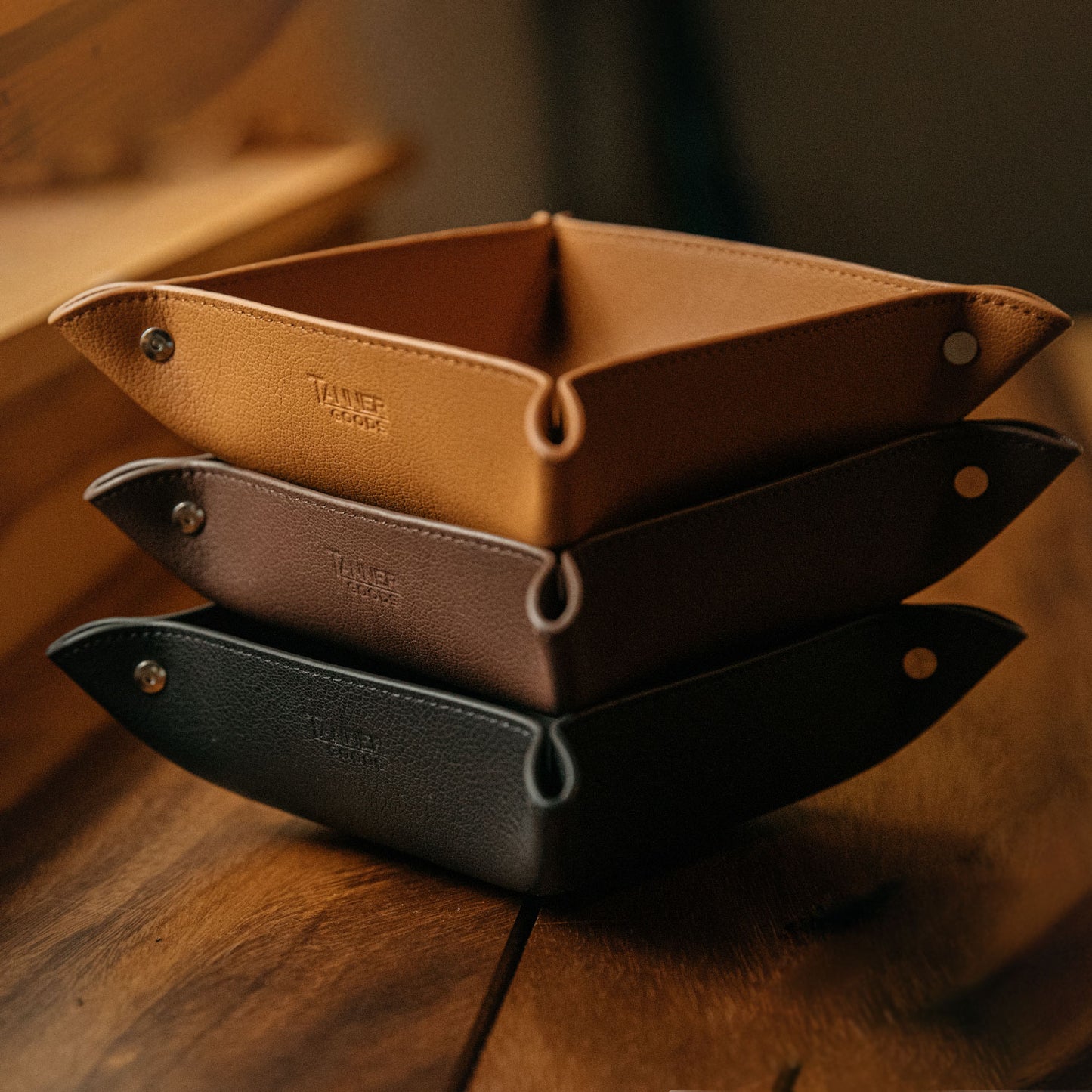 Saddle tan grained recycled leather valet tray, stacked with dark brown and black trays, on a wooden table.