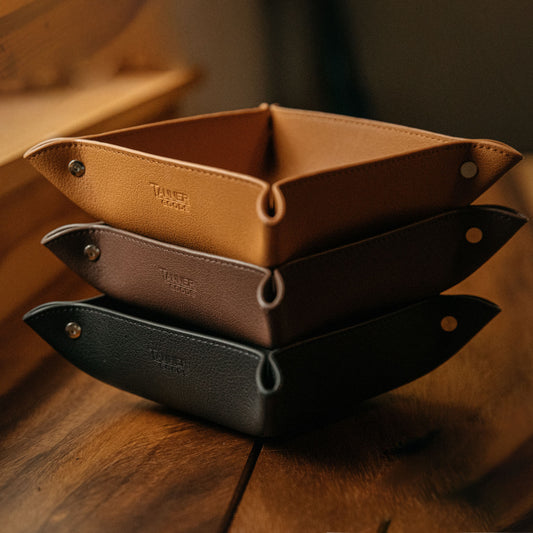 Saddle tan grained recycled leather valet tray, stacked with dark brown and black trays, on a wooden table.