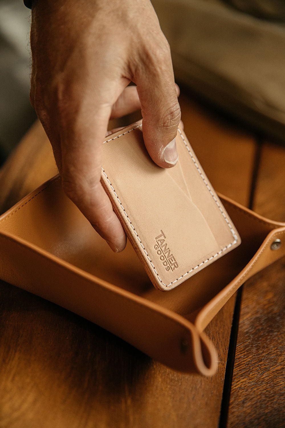 Saddle Tan recycled leather valet tray, a hand placing a natural leather wallet into its folded corners on a wooden surface.