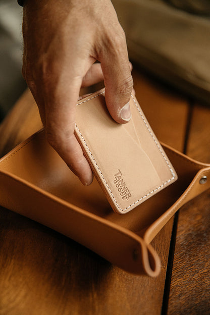 Saddle Tan recycled leather valet tray, a hand placing a natural leather wallet into its folded corners on a wooden surface.