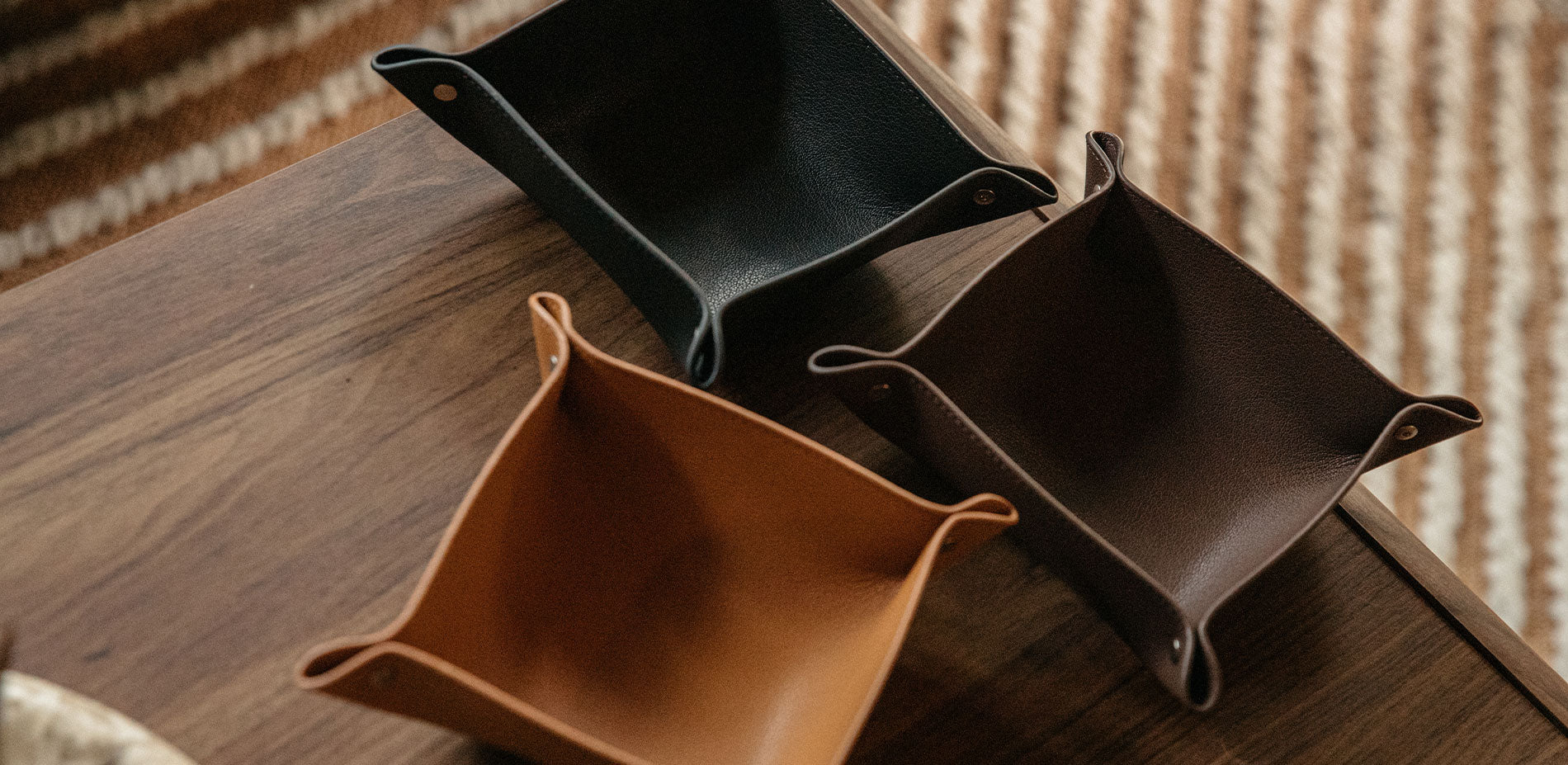 Three leather document trays in tan, dark brown, and black arranged on a wooden surface with striped rug background.