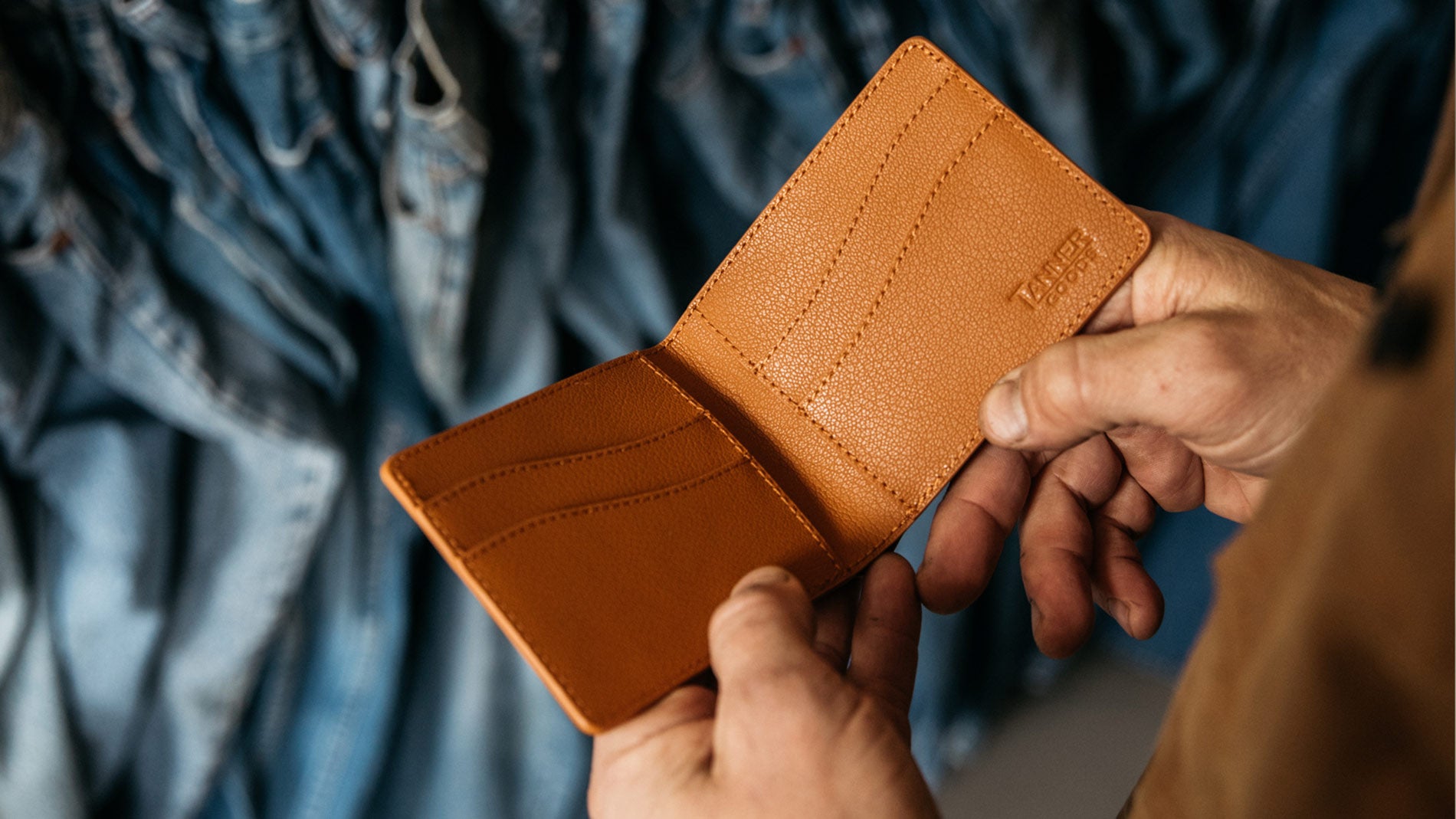 Hands holding open tan leather wallet with card slots, embossed "TANNER GOODS" logo, against blurred blue denim background.