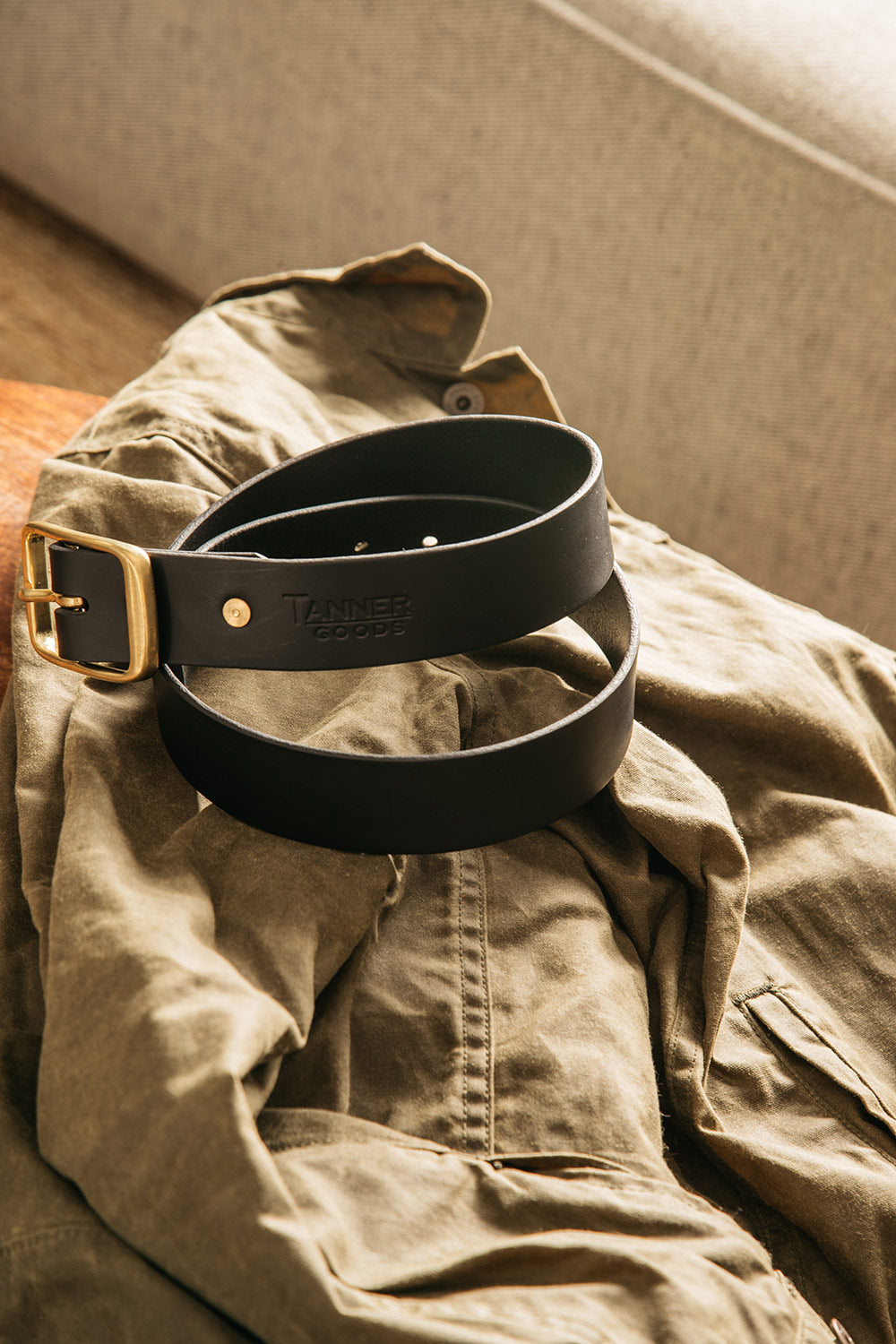 Black leather Standard Belt by Tanner Goods with a brass buckle, coiled on a wrinkled olive green jacket.