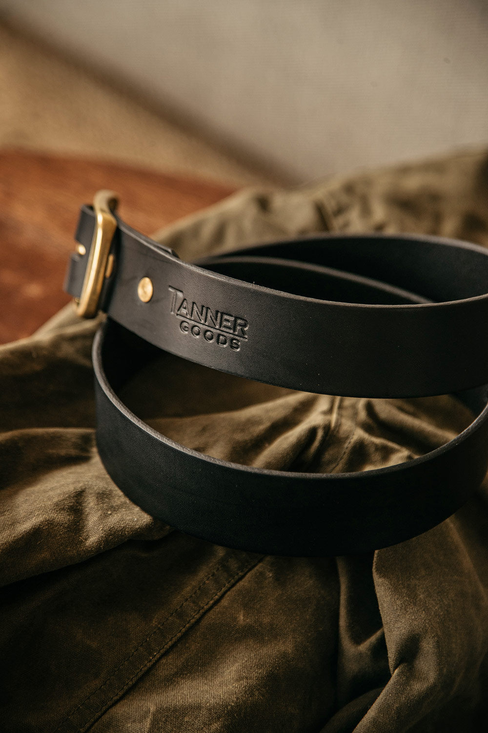Black leather standard belt by Tanner Goods with a brass buckle and gold rivets, coiled on olive green fabric.