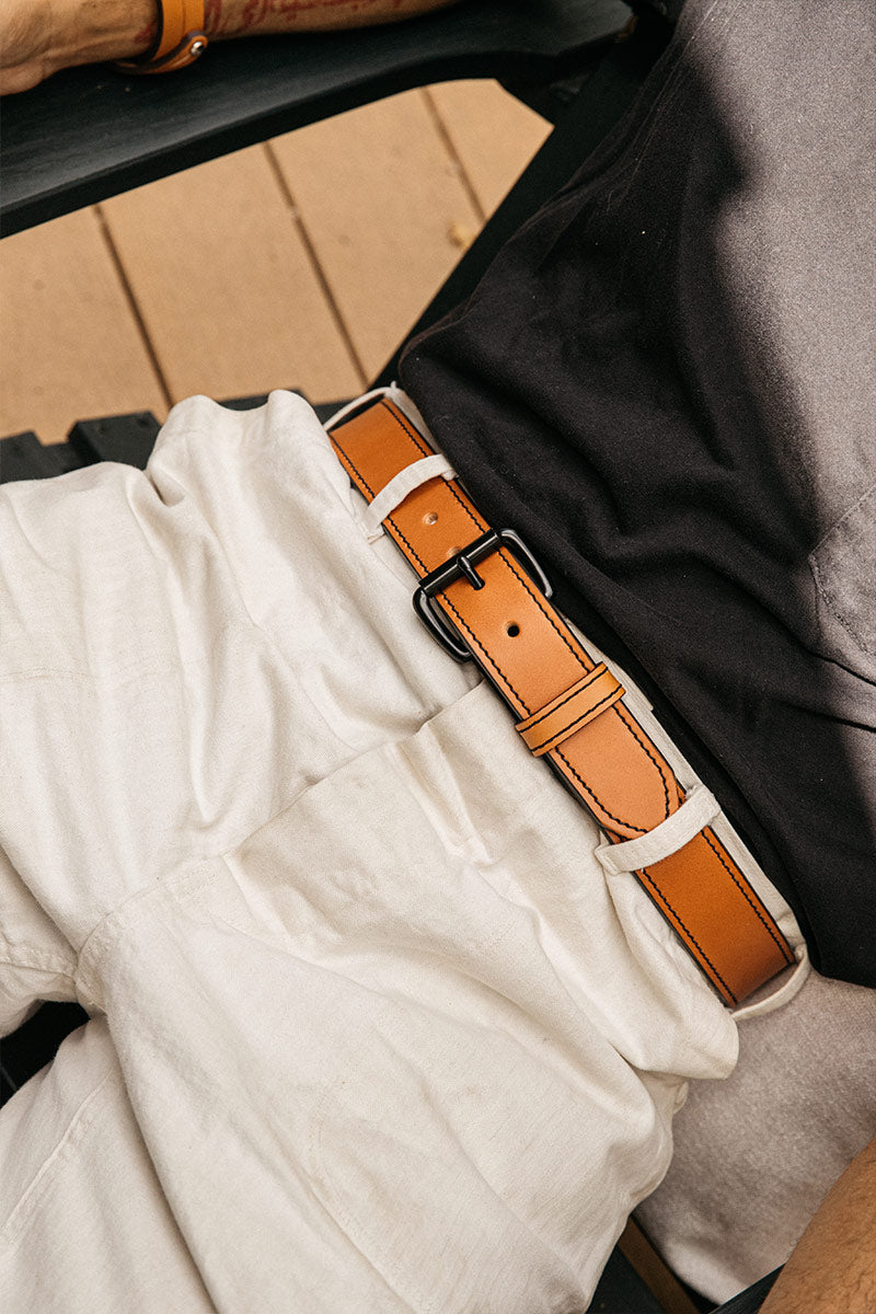 Saddle Tan leather belt with black stitching and a dark buckle, worn with a white linen shirt and gray pants.