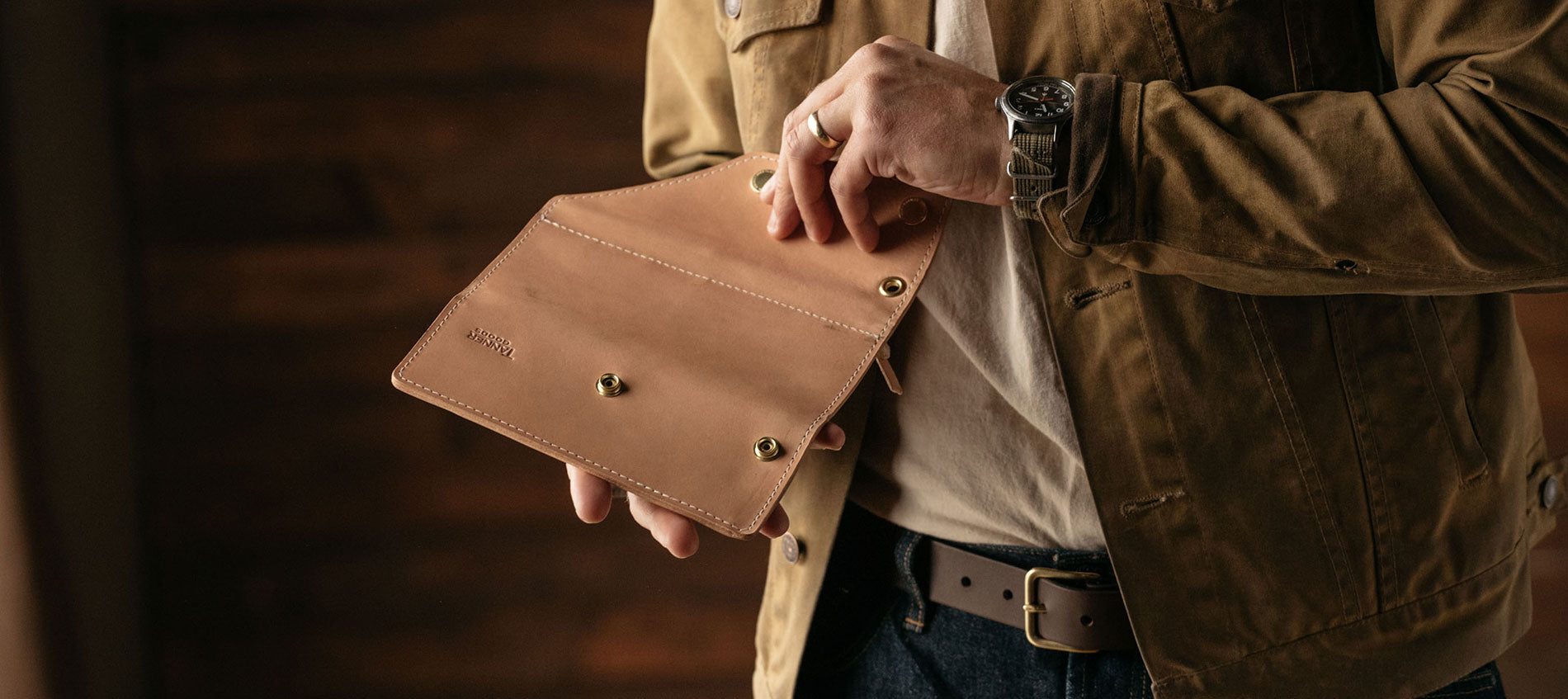 Man in tan jacket holds open tan leather wallet with brass hardware, wearing watch and ring, warm lighting.
