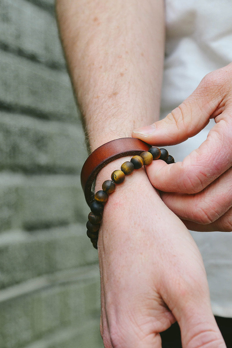 Cognac leather Single Wrap Wristband on a person's wrist, next to a hand adjusting a beaded brown and gold bracelet.