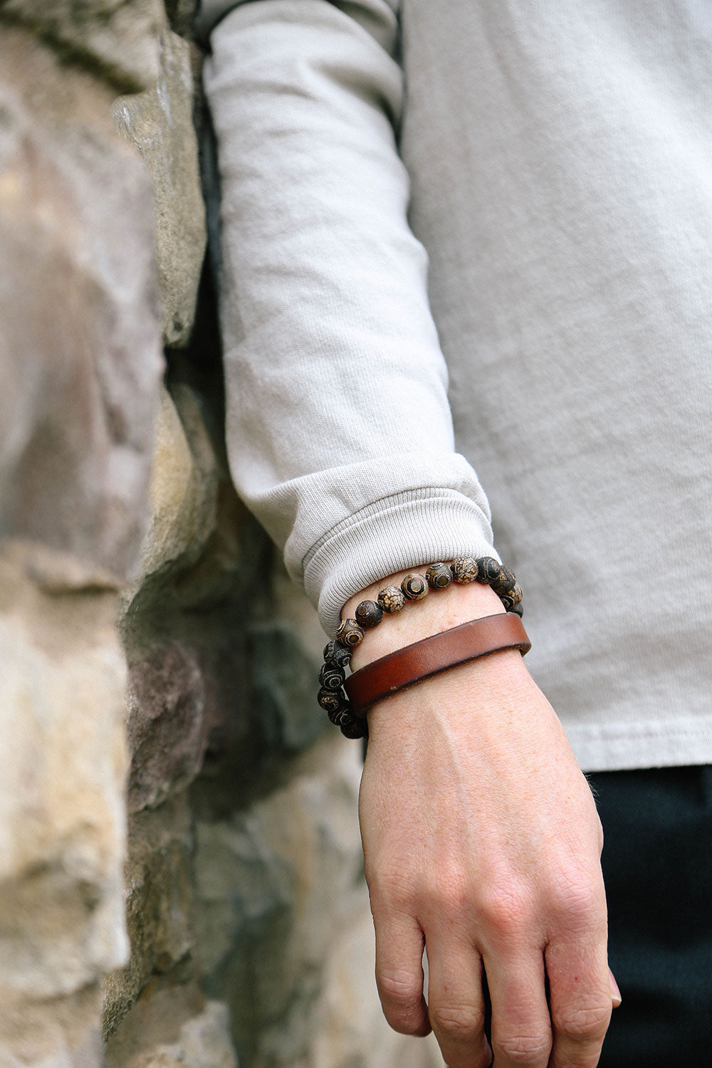 Cognac leather single wrap wristband worn on a wrist, alongside two dark beaded bracelets and a light gray shirt.