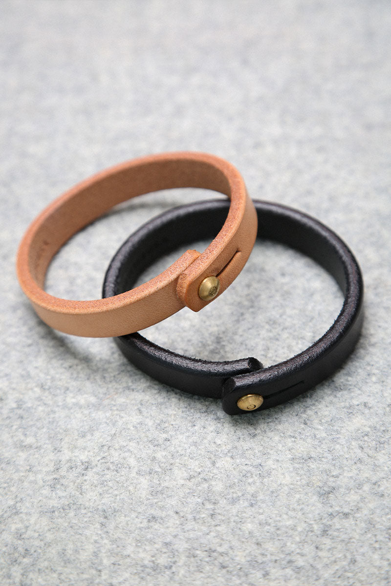 Natural leather single wrap wristband with a brass button stud closure, shown with a black leather wristband on a grey surface.