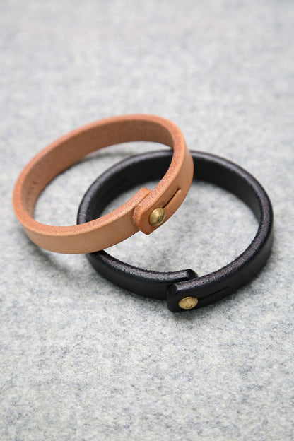 Natural leather single wrap wristband with a brass button stud closure, shown with a black leather wristband on a grey surface.