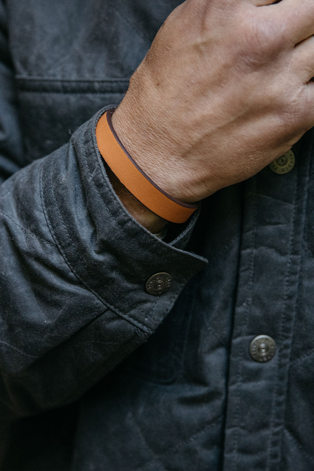 Saddle Tan leather single wrap wristband with dark brown edge, on a wrist against a dark textured jacket with metal buttons.