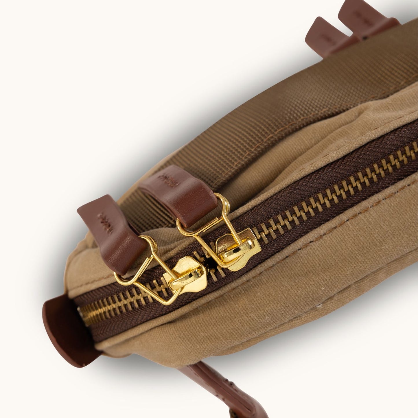 A utility tan waxed canvas mini crossbody bag with brown leather zipper pulls and shiny brass zippers, shown in an angled close-up.