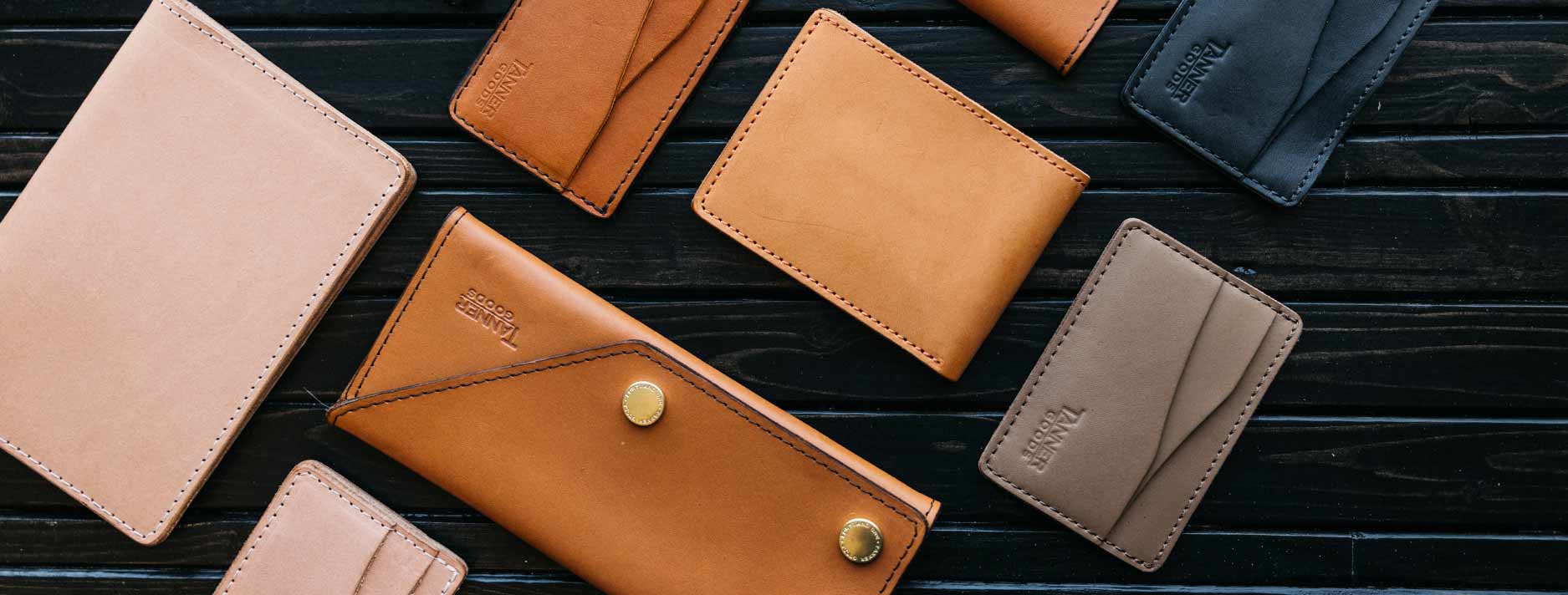 Leather wallets in tan, brown, and black arranged on dark wood, featuring "TANNER GOODS" embossed branding and stitched edges.