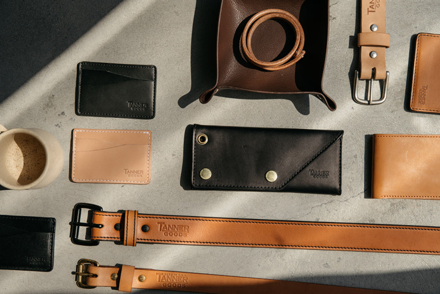 Brown and black leather goods including belts, wallets, and card holders arranged on concrete, with "Tanner Goods" embossed on each item.