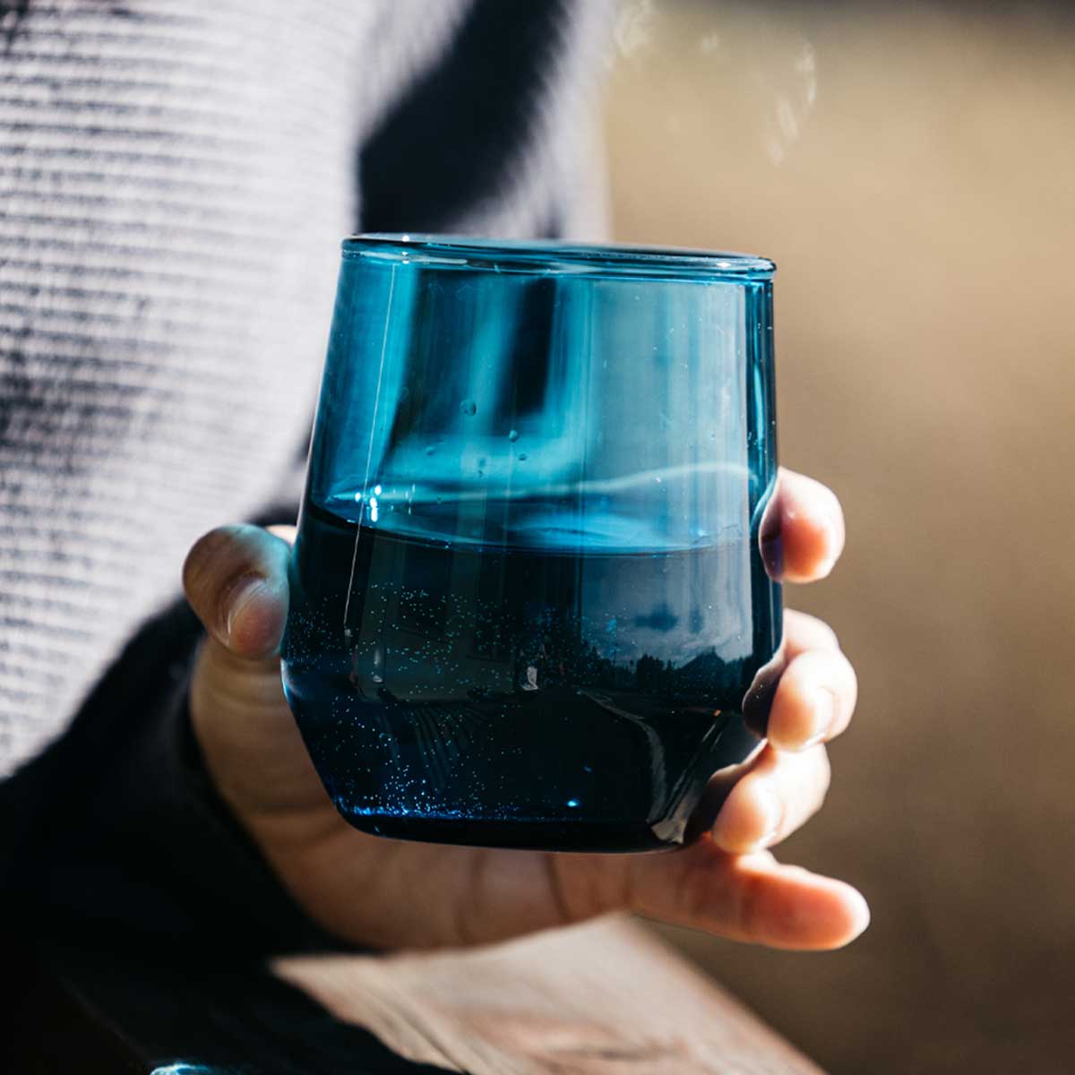 Blue stemless wine glass held by a hand, filled with bubbly clear liquid, against a person in a grey sweater.