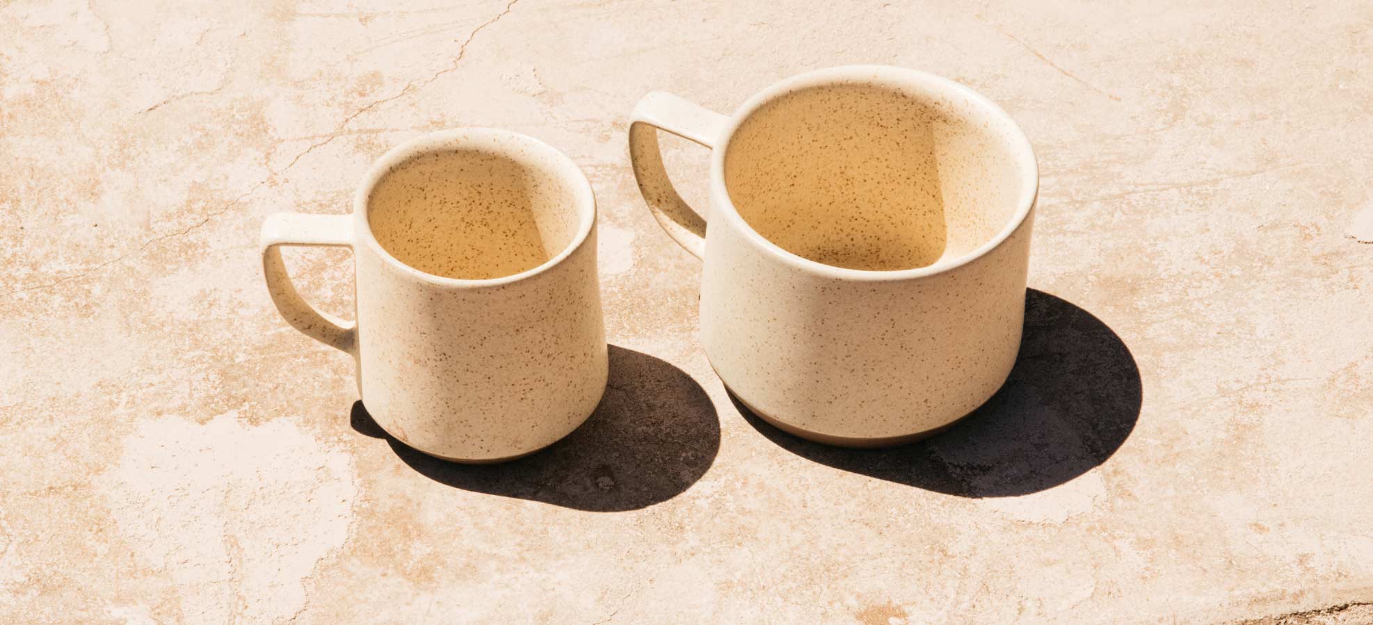 Two beige speckled ceramic mugs filled with coffee sit side by side on a textured stone surface, casting dark shadows.