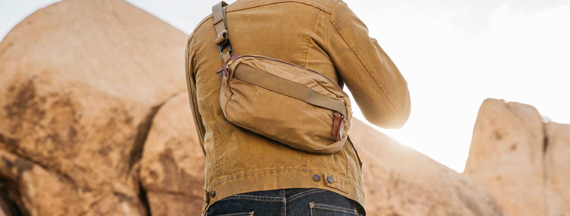 Person in tan jacket and jeans wears matching crossbody bag, standing before large sandstone rocks under bright sky.