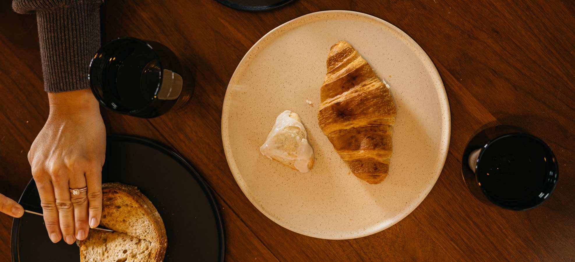 Golden croissant and butter on beige plate, hand slicing bread on dark plate, two black cups on wooden table.