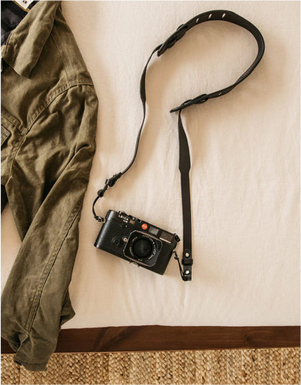 Black camera with leather strap lies on white bedsheet beside olive green jacket, warm natural light.