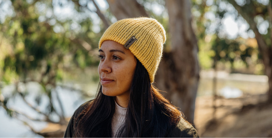 Woman wearing yellow knit beanie with small tag, standing outdoors near trees and water, soft natural light.